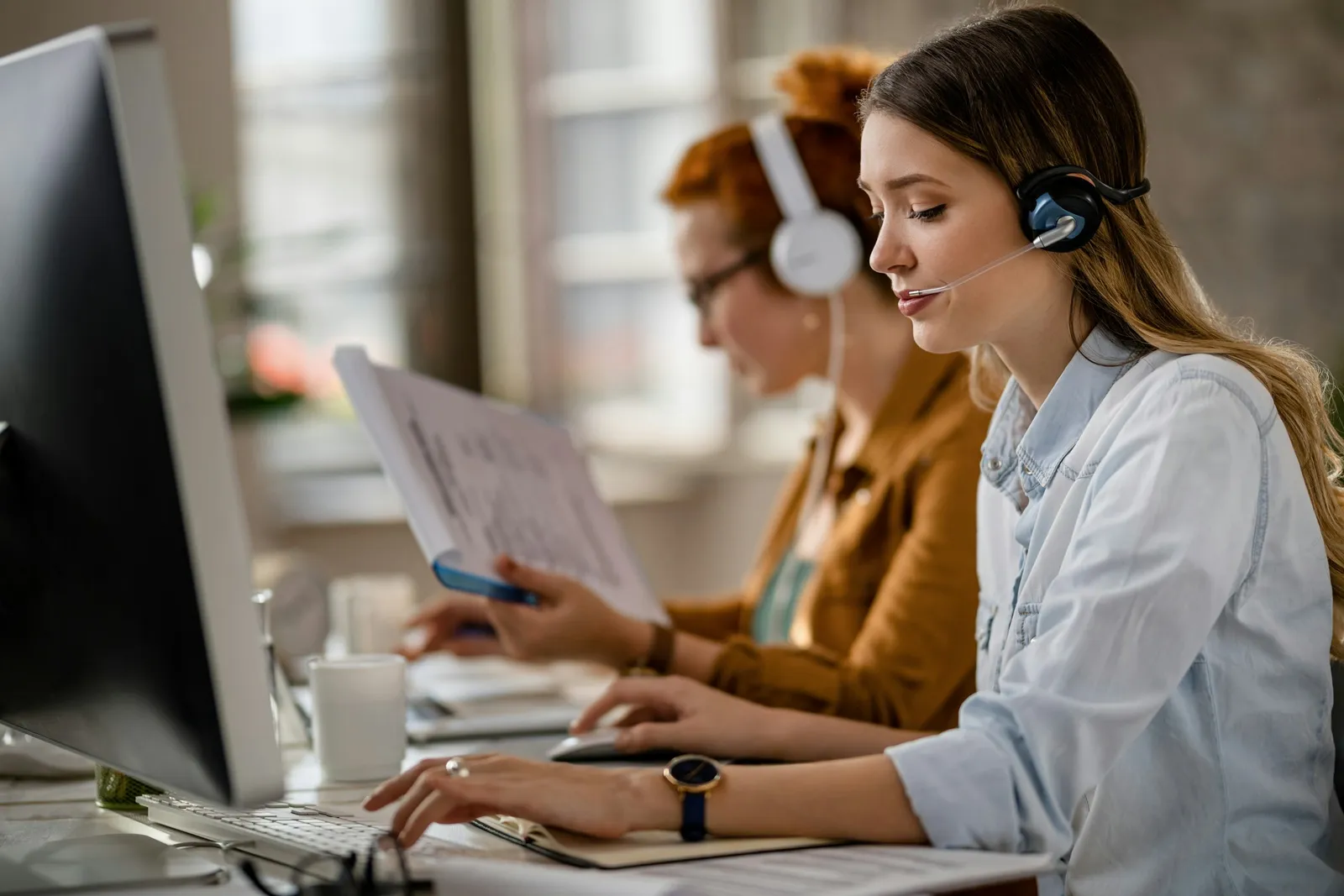 Une jeune secrétaire avec un casque-micro parle en tapant sur son clavier, une collègue travaillant derrière elle.
